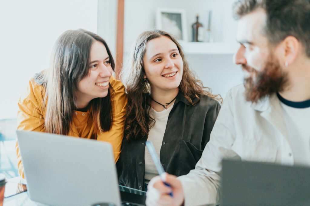 Three Young Adults Engaged In A Business Discussion Around A Laptop In A Bright Room.