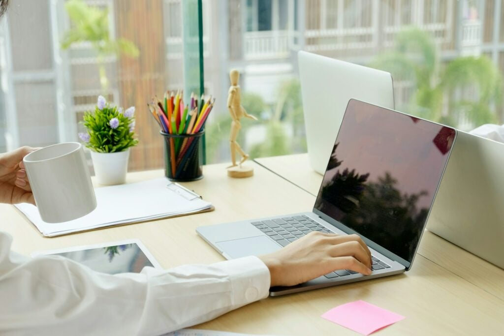 A person types on a laptop with one hand while holding a coffee mug with the other, seamlessly navigating FlowLogic. The desk also has a clipboard, tablet, plant, pencils, and a small wooden mannequin.