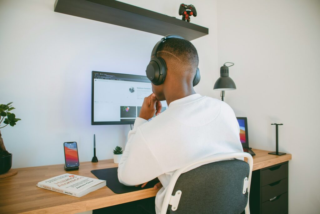 A man wearing headphones is sitting at a desk in his home office, in front of a computer.