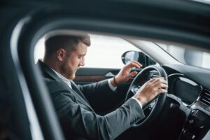 A man in a suit sits in the driver's seat of a car, adjusting the steering wheel with a focused expression, pondering how salary sacrifice could work for his vehicle choice.