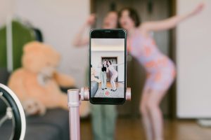 Two people pose in front of a smartphone on a tripod, capturing their image with the perfect backdrop—a large teddy bear. They're crafting content as eye-catching as an Instagram Reel, mindful of how long it can be to hold viewers' attention.