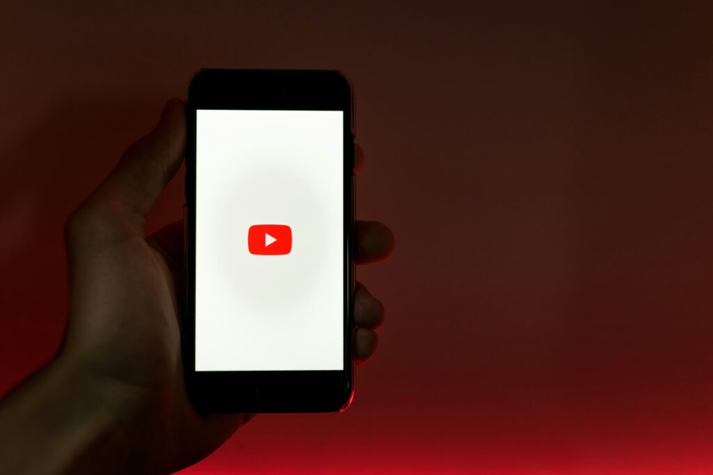 A hand holds a smartphone displaying the YouTube logo on a white screen against a red backdrop, subtly hinting at questions like "How Much Does YouTube Shorts Pay?