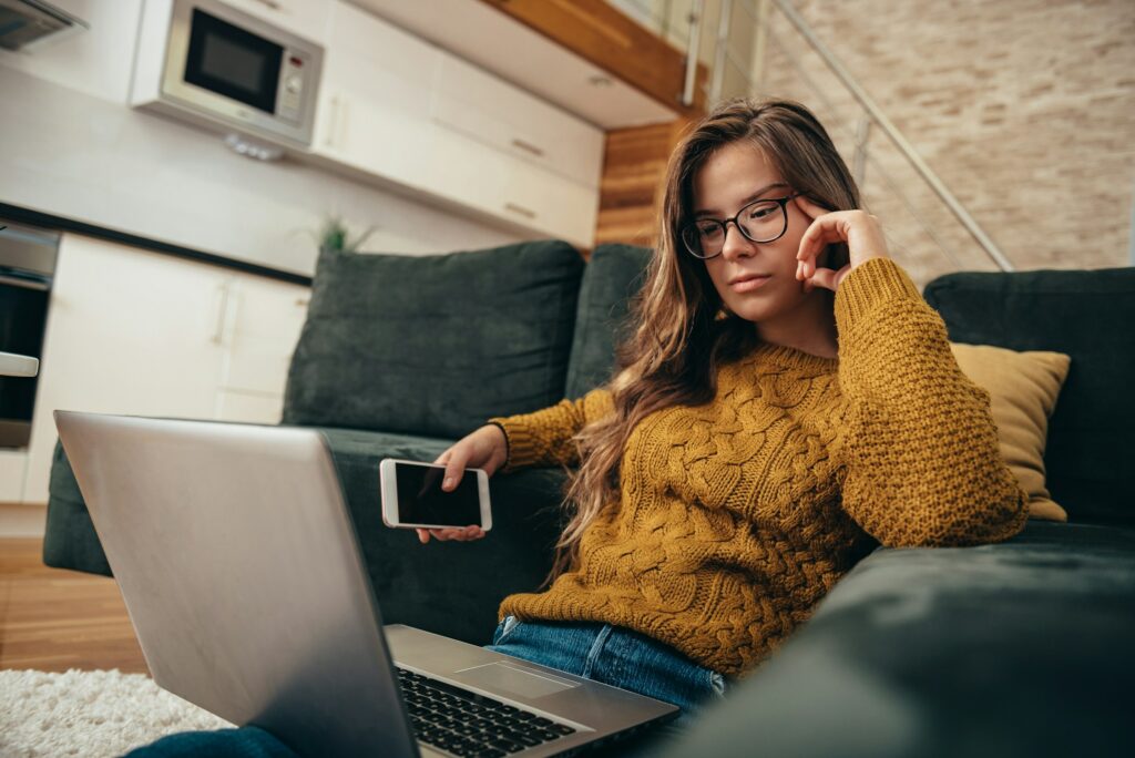 A woman sitting on a couch with a laptop and cell phone, browsing the Ultimate Guide to Add Promotion on LinkedIn 2024.