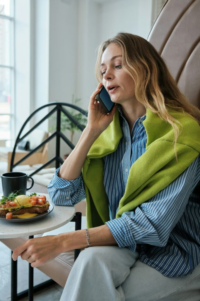 A woman sitting at a table talking on a cell phone, figuring out how to block spam calls on her iPhone.