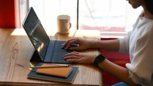 A woman typing on a laptop in a coffee shop, working on how to create a group email list in Outlook.