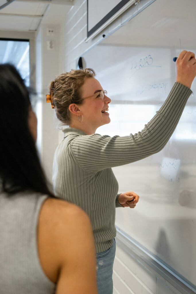 Two women streamlining the process by writing a workflow chart on a whiteboard in a classroom.