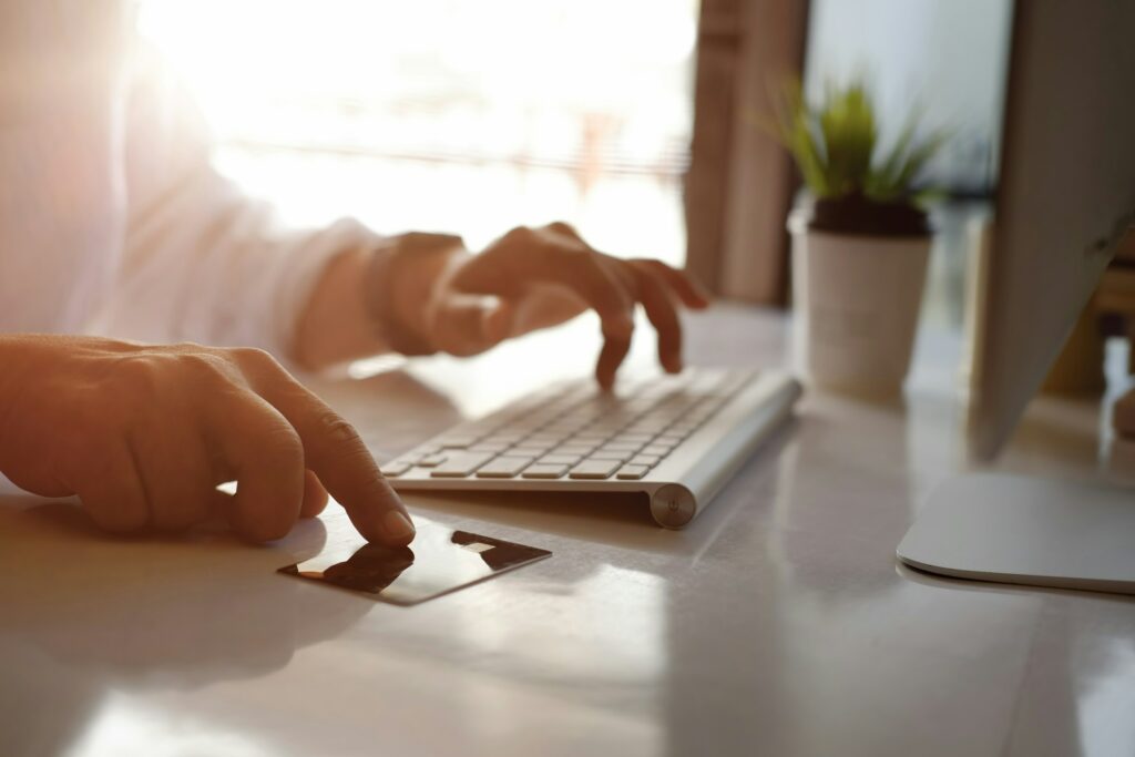 A person typing on a keyboard with a credit card nearby, next to a computer monitor and a plant in the background, searches for "How to Delete Credit Card from Amazon.