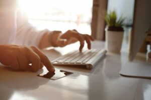 A person typing on a keyboard with a credit card nearby, next to a computer monitor and a plant in the background, searches for "How to Delete Credit Card from Amazon.