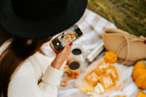 Person in a black hat taking a photo of a picnic setup with snacks, drinks, and a bag on a checkered blanket, possibly looking for inspiration on how to find trending audio on Instagram.