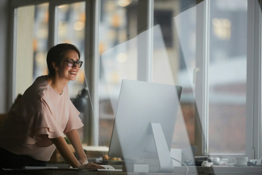A person wearing glasses and a light-colored top leans over a desk, smiling while looking at a large computer monitor in a bright, modern office space, possibly researching how to find the best search keywords per topic on LinkedIn.