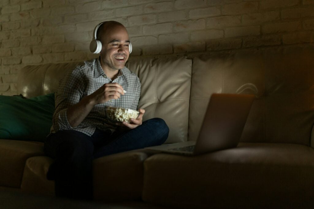 A person wearing headphones sits on a sofa, eating popcorn, and watching a laptop screen in a dimly lit room, perhaps figuring out how to put subtitles on BINGE.