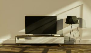 A flat-screen TV sits on a modern TV stand with shelves, next to a floor lamp with a black lampshade, in a sunlit room with shadows from the window. You can enjoy your shows even more if you know how to remove subtitles from your Samsung TV for an unobstructed view.