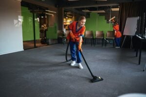 A person in orange and blue uniform expertly vacuums a carpeted room, with chairs neatly stacked against the wall and another individual working diligently in the background—an inspiring snapshot for those wondering how to start a cleaning business.