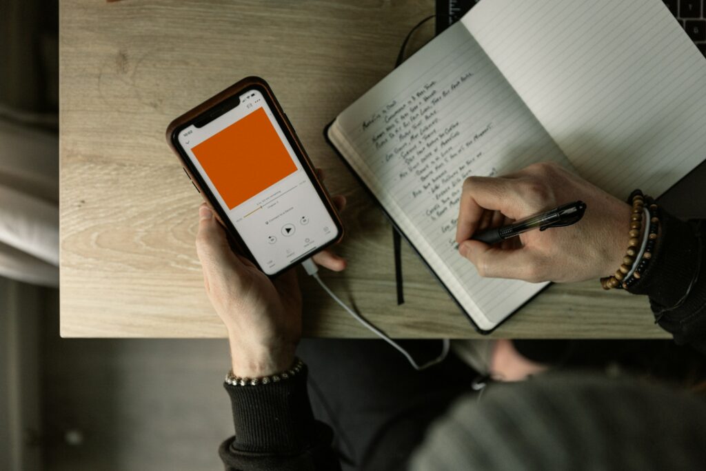 Overhead View Of A Person Holding A Smartphone With A Blank Orange Screen Displaying &Quot;How To Make A Podcast On Spotify&Quot; Next To A Notebook On A Wooden Table.