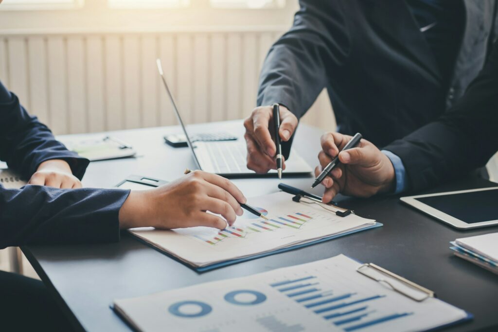Three people in business attire are deep in discussion about the legitimacy of digital marketing, analyzing graphs and charts at a table with pens in hand.