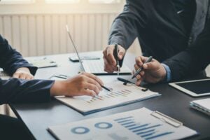 Three people in business attire are deep in discussion about the legitimacy of digital marketing, analyzing graphs and charts at a table with pens in hand.