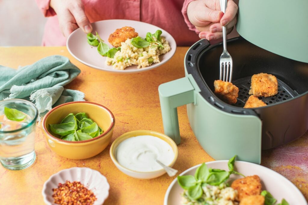 A person uses a fork to transfer breaded food from a green air fryer to a plate with greens and rice. Surrounding dishes include sauce, red pepper flakes, and fresh basil leaves. Is Frigidaire a good brand for kitchen appliances? You might be pleasantly surprised by their reliable performance.