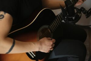 A person with a tattoo on their arm playing an acoustic guitar near a microphone, perhaps pondering, "Is Guyker a good brand?