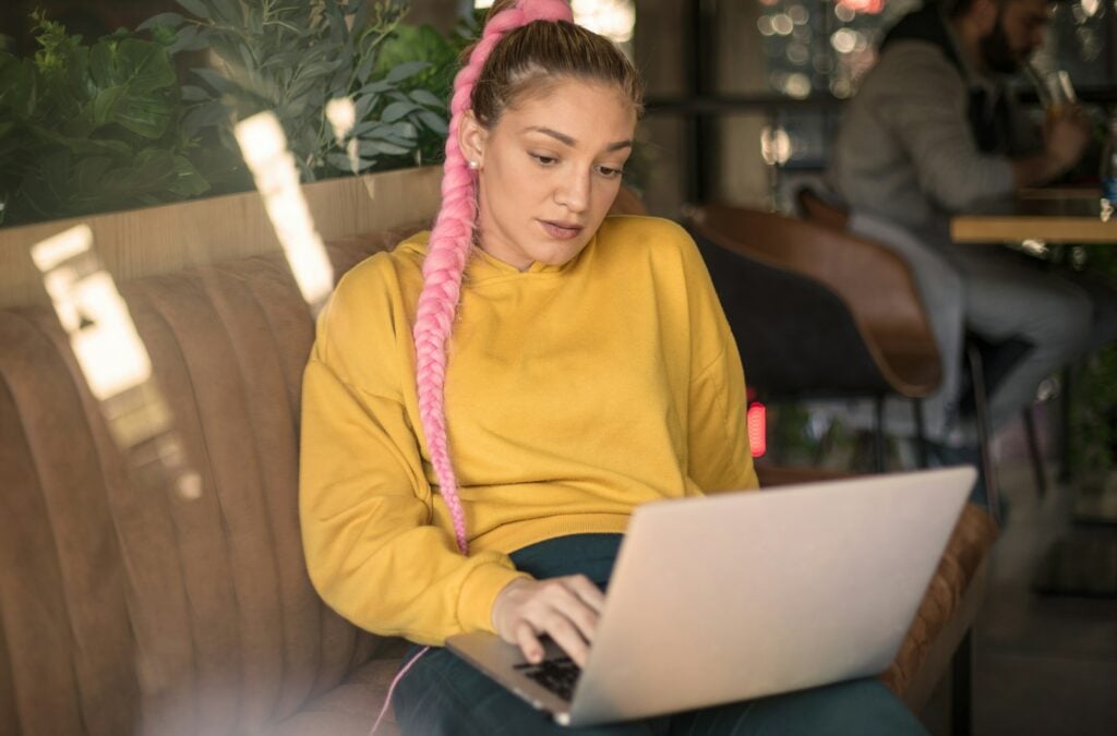 A person with a long pink braid and a yellow hoodie is sitting on a brown sofa in a cafe, focused on their laptop, possibly using imagetotext.io for reliable results.