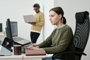 A person sits at a desk typing on a laptop connected to a desktop monitor, with "Is Outlier AI Legit?" displayed onscreen. In the background, another individual stands using a laptop. A mug and notebook rest beside them.