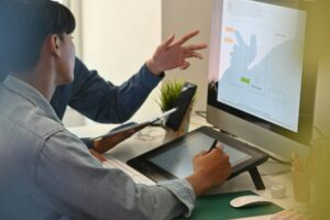 Two people working at a desk with a computer and tablet, discussing SEO strategies for small businesses. One person is pointing at the screen, while the other uses a stylus on the tablet, pondering if investing in SEO is worth it for their growing enterprise.