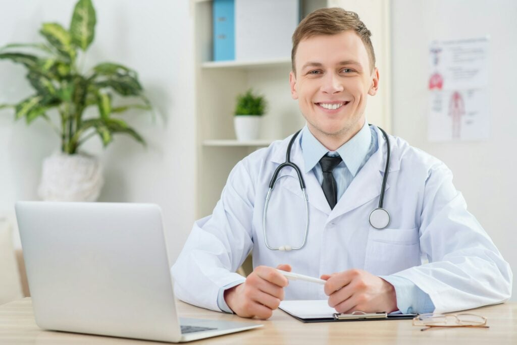 A doctor wearing a white coat and stethoscope sits at a desk with a laptop and plant in the background, smiling at the camera, perhaps pondering, "Is UpDoc Legit?