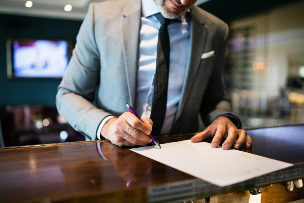 In an office setting, where questions like "Is eDreams legit?" may arise over casual coffee breaks, a man in a gray suit diligently signs a document with a purple pen at the wooden counter.