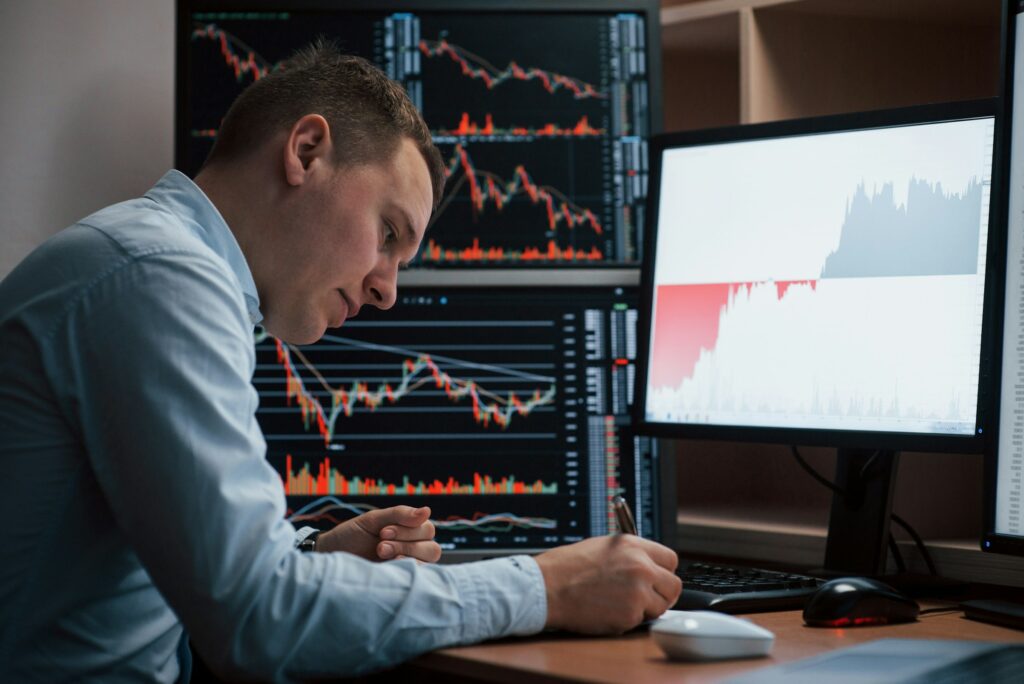 A Man Sitting In Front Of Two Monitors Analyzing Stock Charts, Hoping To Achieve Financial Freedom Through Passive Income Ideas.