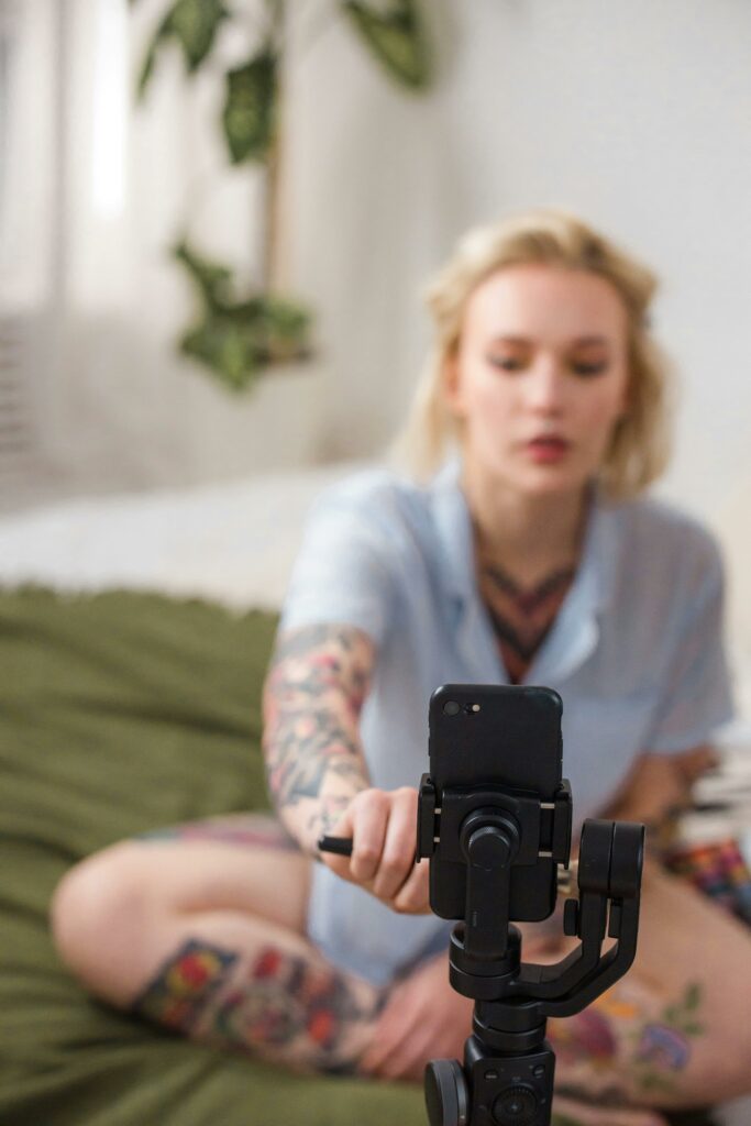 A Woman With Tattoos Sitting On A Bed, Browsing Her Cell Phone And Contemplating Ideas For Achieving Financial Freedom Through Passive Income.