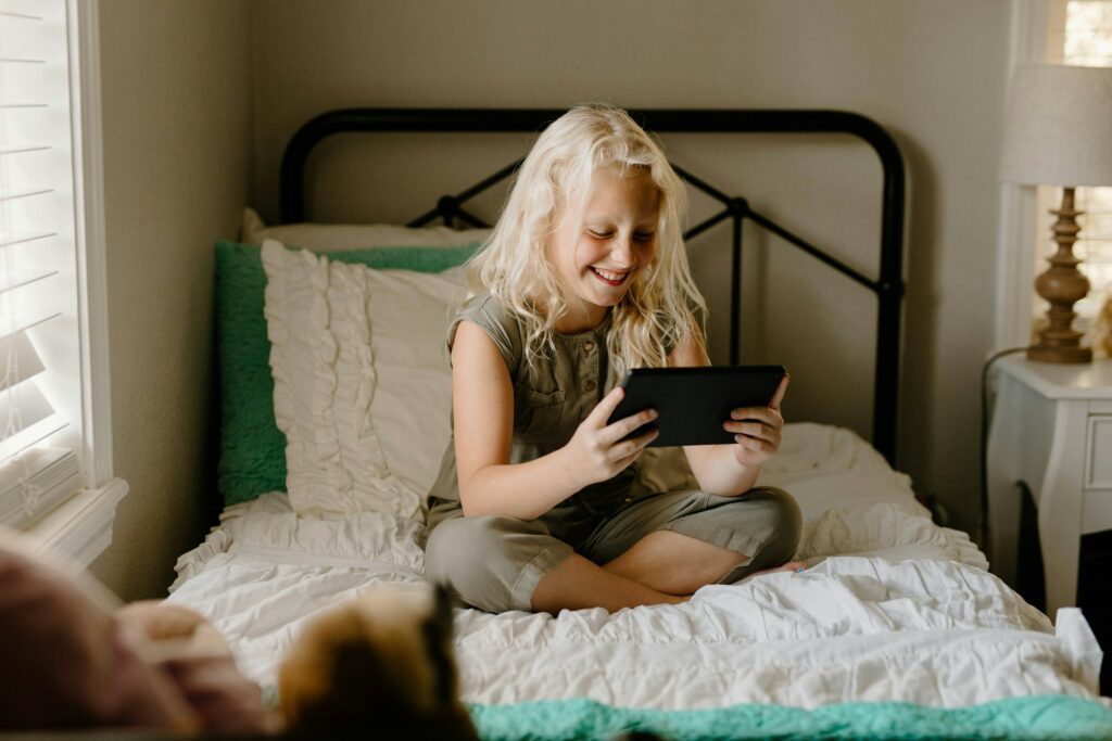 A young girl with blonde hair sits cross-legged on a bed, smiling as she enjoys some screen time on her tablet.