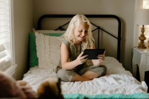 A young girl with blonde hair sits cross-legged on a bed, smiling as she enjoys some screen time on her tablet.