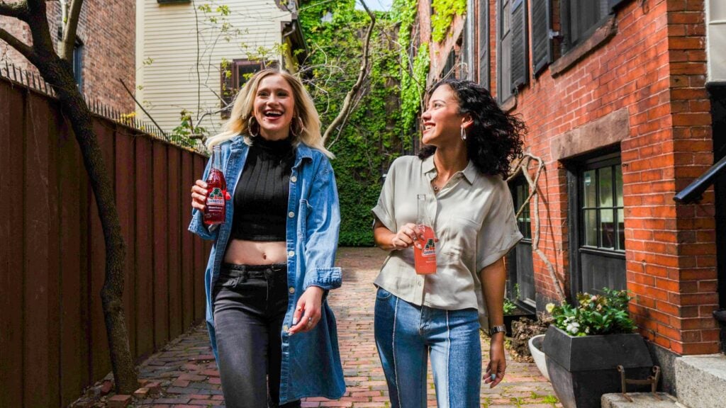 Two women, discussing strategies for 'What Is Field Marketing,' walk and smile along a brick pathway between buildings, holding bottled drinks.