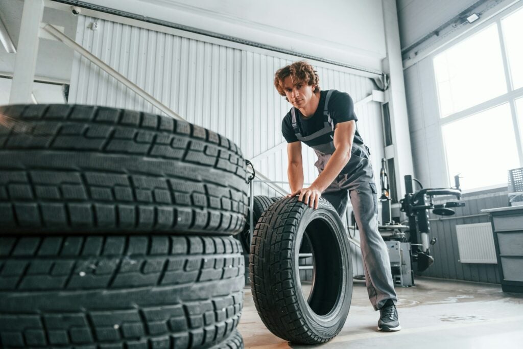 A worker rolls a tire past a stack of tires inside an automotive workshop under natural light and a high ceiling, pondering what is the best tire brand for optimal performance.