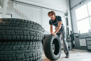 A worker rolls a tire past a stack of tires inside an automotive workshop under natural light and a high ceiling, pondering what is the best tire brand for optimal performance.