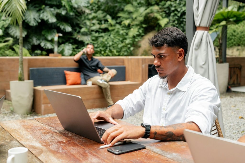 A man in a white shirt is researching "What are Orphan Pages" on his laptop at a wooden table outdoors, surrounded by lush greenery. In the distance, another person sits quietly, enjoying the serene environment.