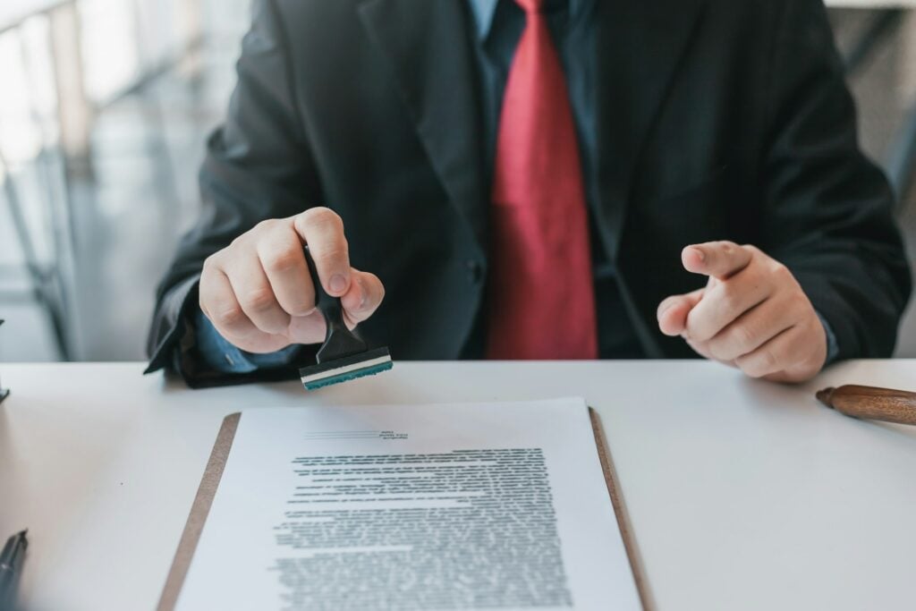 A person in a suit, processing an environmental compliance certificate, stamps a document on a clipboard, holding a rubber stamp above the desk.