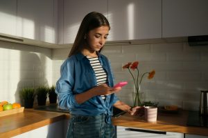 A woman in a kitchen holds a mug while looking at a smartphone, perhaps researching "What is Google Workspace." A counter with fruit, plants, and flowers decorates the background as sunlight casts shadows in the room.