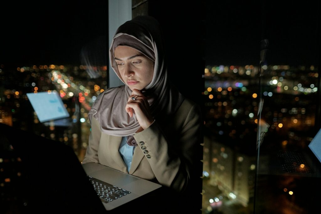A Woman In A Hijab Is Using A Laptop At Night To Fix Keyword Cannibalization Through Simple Steps.