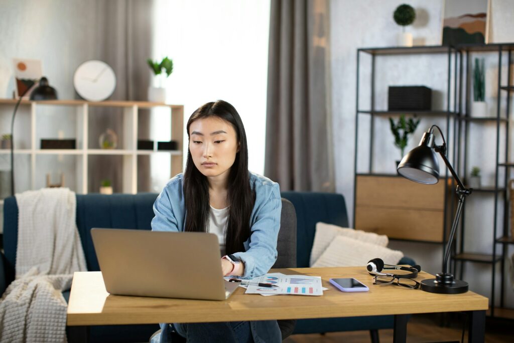 A person sits at a table drafting a mid-cycle marketing email on their laptop, surrounded by papers, a phone, and a microphone in a modern living room.
