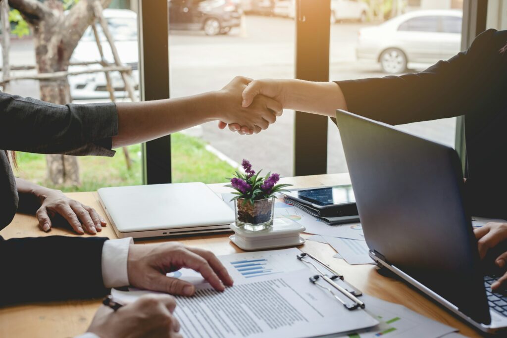 Two people shaking hands across a table with a laptop, documents, and a small potted plant. Another person is holding a pen and reading a document titled "What is a Share Purchase Agreement.