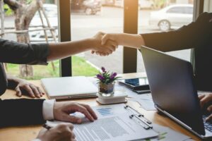 Two people shaking hands across a table with a laptop, documents, and a small potted plant. Another person is holding a pen and reading a document titled "What is a Share Purchase Agreement.