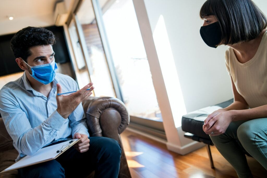 An Allied Health Professional wearing a surgical mask provides care to a woman sitting on a couch in the healthcare system.