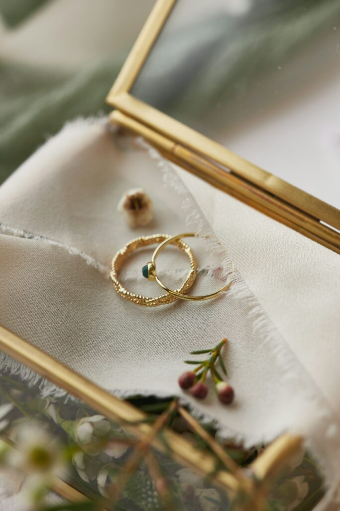 A pair of gold rings and a small earring rest on a piece of fabric inside a gold-framed glass box, evoking questions like, "What is the oldest jewelry brand?" Nearby, a small sprig with red berries adds a touch of nature.