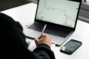 Person with pen taking notes in front of a laptop displaying a financial chart, contemplating, "What is the option wheel strategy?" with a smartphone beside them on the desk.