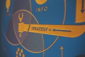 Close-up of a blue and orange graphic showing interconnected concepts. A hand holding a wrench points to the word "Strategy" with arrows, alongside the word "Info" in the background.