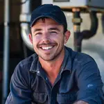 A man in a dark cap and blue shirt is smiling while sitting in front of industrial equipment.