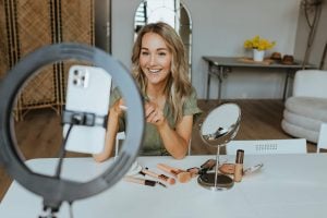 A smiling woman sits at a table with makeup tools and a ring light, recording a video on her phone, aiming to reach a wider audience by using CapCut Auto Captions for enhanced accessibility.