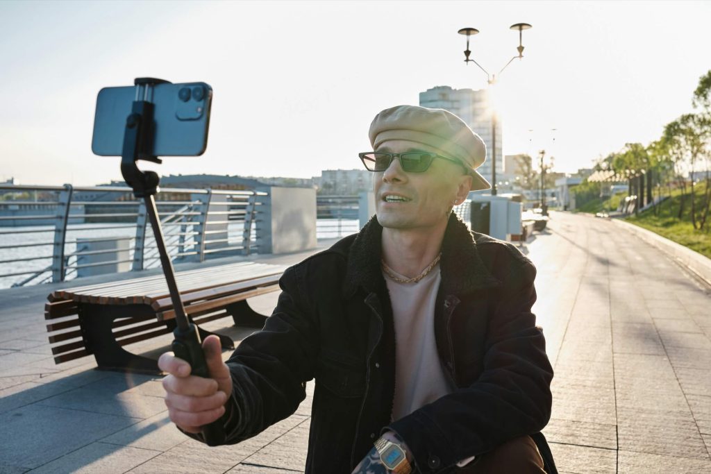 A person wearing sunglasses and a beret takes a selfie with a smartphone on a stick, sitting on a riverside promenade with benches and lampposts in the background.