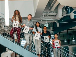 A group of five people are standing on a staircase, holding individual letters that together spell out "BRAND." They are smiling and casually dressed in a modern office setting.