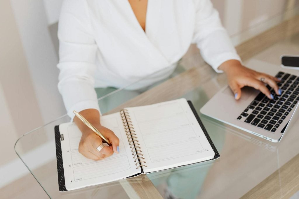 Person in white shirt writes in a planner with a gold pen while using a laptop on a glass table.